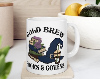 May include: White ceramic mug with a black and blue illustration of a witch's hat with books and a cold brew coffee cup. The text "Cold Brew Books & Covens" is printed on the mug.