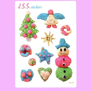 May include: A sheet of stickers featuring various Christmas-themed designs made from playdough. The designs include a Christmas tree, a star, a snowman, an angel, a heart, a snowflake, and a donut. The text "J.S.S. stickers" is at the top of the sheet.