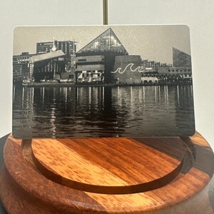 May include: A grayscale image of a cityscape, likely a souvenir or collectible card, featuring a waterfront scene with buildings and reflections in the water. The card is displayed on a wooden stand.