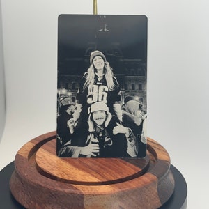 May include: A black and white metal photo card featuring a group of people. A person wearing a jersey with the number 36 is sitting on another person's shoulders. The card is displayed on a wooden stand.