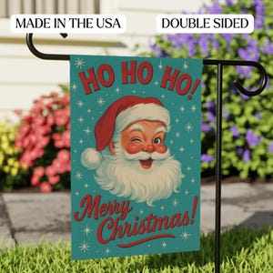 Peut inclure: Un drapeau de jardin recto-verso avec le Père Noël qui cligne de l'œil, avec les mots "Ho Ho Ho!" et "Merry Christmas!" en rouge sur un fond turquoise. Le drapeau est affiché sur un support en métal noir dans un jardin.