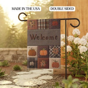 May include: Double-sided garden flag with a patchwork design featuring pumpkins, flowers, and plaid patterns. The word "Welcome" is embroidered on a gray panel. The flag is displayed on a black metal stand, perfect for fall decor.