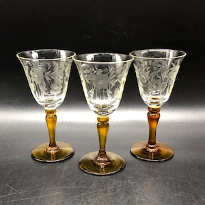 May include: Three clear glass cordial glasses with etched floral designs. The stems and bases are amber-colored. The glasses are arranged in a row against a dark background. The glasses are likely vintage or antique.