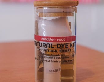 May include: A clear glass jar with a wooden lid contains a natural dye kit. The label reads "madder root NATURAL DYE KIT for NATURAL FIBERS." Inside are small containers and packets of dye extract, mordant, and soda ash. The kit is designed for natural fibers.