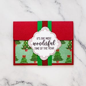 Puede incluir: Un tarjetero navideño rojo con una banda verde y roja con purpurina. El tarjetero tiene un frente estampado con árboles de Navidad y el texto "IT'S THE MOST WONDERFUL TIME OF THE YEAR" en una etiqueta blanca festoneada.