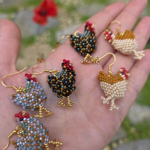 May include: A collection of beaded chicken earrings in various colors, including black, gold, white, and blue. Each earring features a red comb and gold-colored metal hooks and feet. The earrings are displayed on a hand against a blurred green background.