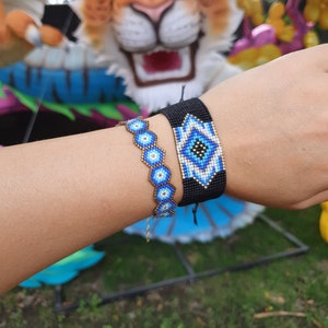 Op de afbeelding: Twee kralenarmbanden met blauwe en witte oogontwerpen op een zwarte achtergrond. De armbanden worden om de pols gedragen.