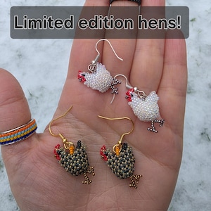 May include: A hand displays beaded jewellery, including a black cat ring, two pairs of chicken earrings, and a colourful ring. The earrings feature white and black chickens with red accents. The text "Limited edition hens!" is displayed.