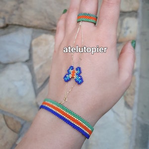 Könnte beinhalten: Ein goldenes Kettenarmband mit einem blauen und orangefarbenen Schmetterlingsanhänger. Das Armband ist mit einem Ring mit grünen und orangefarbenen Bändern verbunden. Der Text "atelutopier" ist im Bild sichtbar.