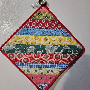 May include: A red, diamond-shaped pot holder with a patchwork design. The pot holder is made of different fabrics with floral and geometric patterns in shades of red, blue, yellow, green, and pink.