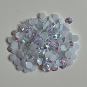 May include: A collection of white, iridescent rhinestones. The rhinestones are circular with a flat reverse. The iridescent stones reflect a spectrum of colours, including pink, purple, and green. The rhinestones are scattered on a white surface.