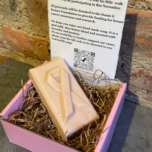 Puede incluir: Una caja rosa con papel triturado marrón en su interior. Dentro de la caja hay una barra de jabón rosa y blanca con un diseño de cinta rosa. El jabón es una recaudación de fondos para la Fundación Susan G Komen para proporcionar financiación para la concienciación sobre el cáncer de mama y la investigación. Se incluye un código QR en la tarjeta para donar a la causa.