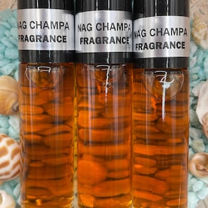 Pu&ograve; includere: Tre bottiglie di vetro ambrato con tappi neri ed etichette argentate che recitano "NAG CHAMPA FRAGRANCE". Le bottiglie sono su una superficie blu e bianca.