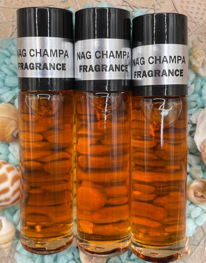 Pu&ograve; includere: Tre bottiglie di vetro ambrato con tappi neri ed etichette argentate che recitano "NAG CHAMPA FRAGRANCE". Le bottiglie sono su una superficie blu e bianca.