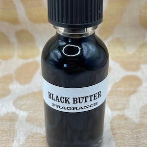 May include: A small glass bottle with a black cap and a white label that reads "BLACK BUTTER FRAGRANCE". The bottle is filled with a dark liquid.