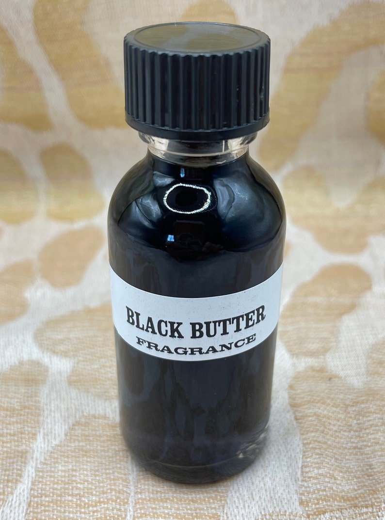 May include: A small glass bottle with a black cap and a white label that reads "BLACK BUTTER FRAGRANCE". The bottle is filled with a dark liquid.