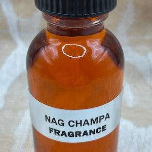 Pu&ograve; includere: Una bottiglia di vetro trasparente con un tappo a vite nero riempita di un liquido marrone. La bottiglia ha un'etichetta bianca con il testo "NAG CHAMPA FRAGRANCE".