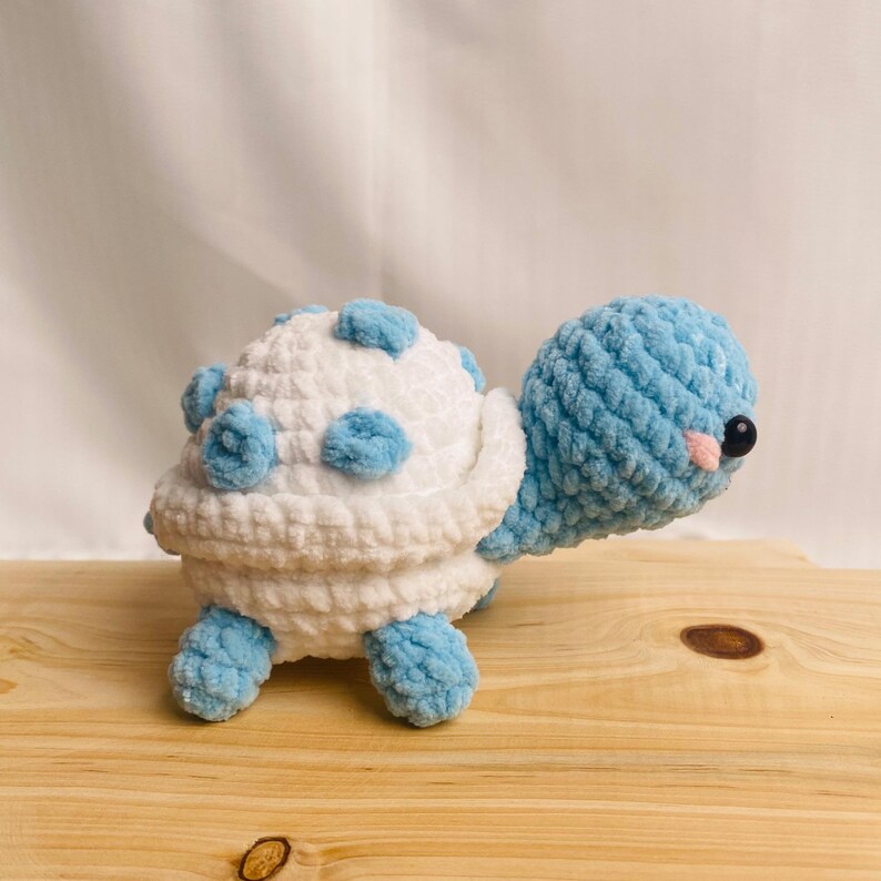 May include: A crocheted turtle plush toy featuring a white shell with light blue spots, a light blue head, and light blue limbs. The turtle has small black plastic eyes and tiny pink stitched cheeks, crafted from soft, textured yarn material.