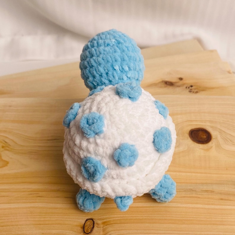 May include: A plush turtle toy crafted from soft, chunky yarn. The turtle features a rounded white shell decorated with several small, light blue circular patches. The head and four small feet are made of the same light blue yarn as the patches.