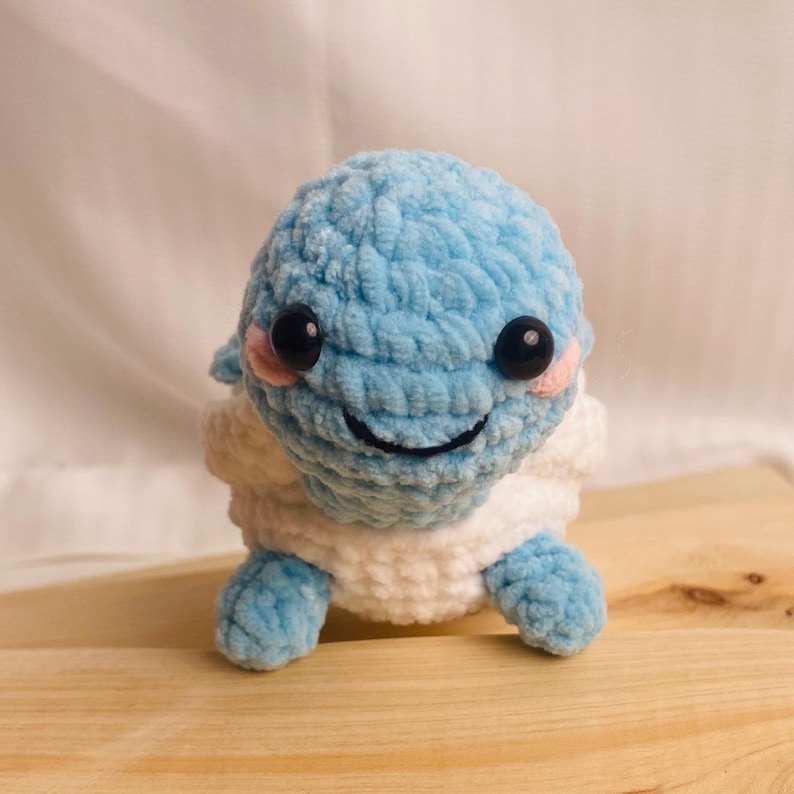 May include: A small, handmade crochet plush toy shaped like a turtle with a light blue head and limbs, a white shell, and two large black plastic eyes. The creature features small pink embroidered cheeks and a simple black stitched smile on its face.