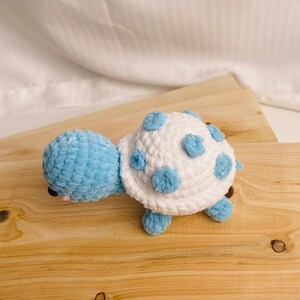 May include: A handmade crochet turtle toy featuring a light blue head and limbs with a white shell adorned with circular light blue spots. The plush toy is crafted from soft yarn and has small black eyes and a tiny pink stitched mouth detail.