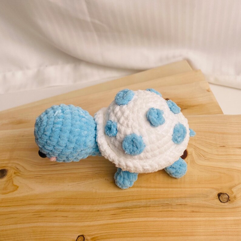 May include: A handmade crochet turtle toy featuring a light blue head and limbs with a white shell adorned with circular light blue spots. The plush toy is crafted from soft yarn and has small black eyes and a tiny pink stitched mouth detail.