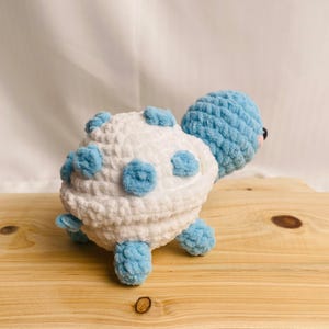 May include: A plush turtle toy crafted from soft, chunky yarn. The shell is white with light blue circular spots, while the head and four small legs are solid light blue. The turtle features a rounded head with a small pink accent near the eye.