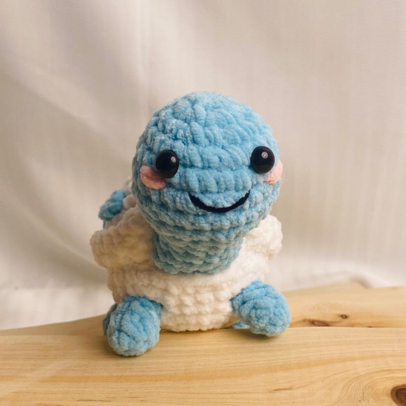 May include: A small, handmade crochet turtle plush toy featuring a light blue head and limbs with a white shell. The turtle has two round black plastic eyes, a stitched black mouth, and small pink circular patches on its cheeks for a blush effect.