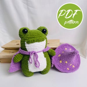 Könnte beinhalten: Ein gehäkeltes Frosch-Stofftier in Grün und Weiß, das einen violetten Umhang mit Bindeband trägt. Neben dem Frosch liegt ein passender violetter Spitzhut, der mit kleinen gelben Sternen verziert ist. Ein grüner Kreis mit dem Text PDF pattern ist zu sehen.
