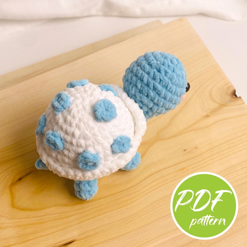 May include: A crocheted turtle plush toy featuring a white shell with light blue spots and a matching light blue head and limbs. The toy is made from soft, textured yarn and includes a circular green graphic with the text PDF pattern.