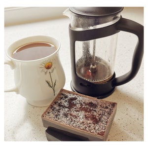 May include: A white ceramic mug with a daisy design filled with coffee, a black French press coffee maker, and a brown coffee-scented soap bar.