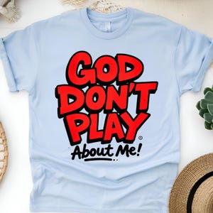 May include: Light blue t-shirt with the text "GOD DON'T PLAY About Me!" in red, outlined in black. The shirt is made of a soft material and has rolled-up sleeves. A straw hat and succulent plant are visible in the background.