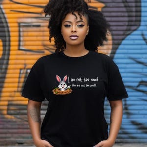 Puede incluir: Camiseta negra con un diseño de conejo de dibujos animados y el texto "I am not too much, You are just too small." El conejo es gris y blanco. La camiseta es una prenda casual.