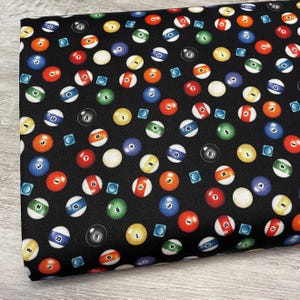 May include: Black fabric featuring a repeating pattern of colorful billiard balls and small blue squares. The balls are numbered 1 through 15, with various colors including red, yellow, green, and blue. The fabric is suitable for various crafting projects.