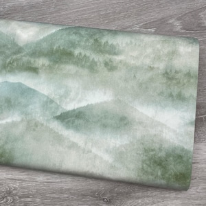 May include: A folded fabric with a watercolor mountain scene. The design features layers of green and gray mountains with a misty, atmospheric effect. The fabric has a soft, muted color palette.