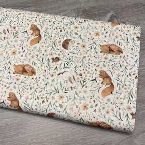 May include: Fabric featuring a repeating pattern of woodland creatures and floral elements. The design includes squirrels, deer, rabbits, hedgehogs, and birds amongst delicate flowers and foliage in brown, orange, and green on a cream background.