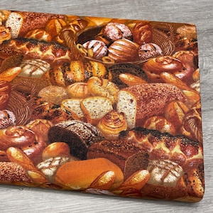 May include: A fabric with a detailed print of various breads, including loaves, rolls, and pastries. The image features a variety of baked goods in shades of brown and tan, with some items in a basket. The overall theme is a bakery or food-related.