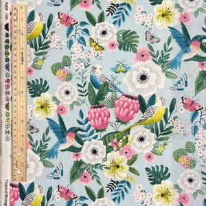 May include: Light blue fabric with a floral and bird pattern. The design features white, pink, and yellow flowers, along with hummingbirds, butterflies, and parakeets. The text "Tropical Menag" is visible.