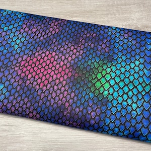 May include: A piece of fabric with a repeating scale pattern in shades of blue, purple, pink, and green. The scales are arranged in a grid-like fashion, creating a textured appearance. The fabric is likely used for crafting or apparel.