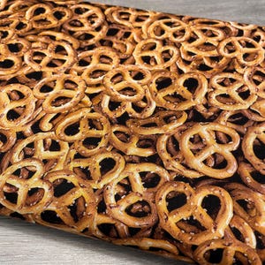 May include: A fabric featuring a repeating pattern of golden-brown pretzels. The pretzels are detailed with visible salt and a dark brown background. The image is a close-up, showing the texture and arrangement of the baked goods.