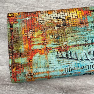 May include: A rolled piece of fabric with a distressed, abstract design. The pattern features a mix of orange, yellow, and teal colors, with hints of brickwork and text. The overall aesthetic is vintage and artistic.