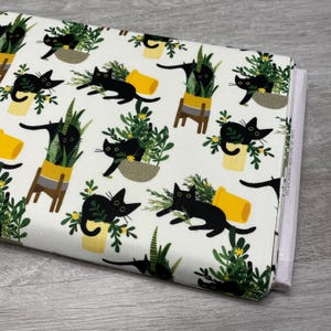 May include: A roll of white fabric featuring a repeating pattern of black cats, potted plants, and yellow pots. The cats are in various poses, and the plants have green leaves and yellow flowers. The fabric is folded and ready for use.