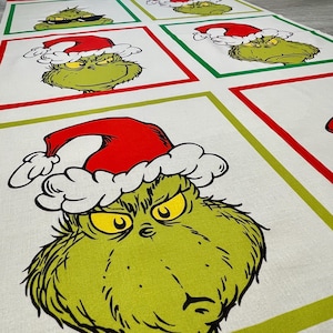 May include: A white fabric table runner featuring a repeating pattern of the Grinch wearing a Santa hat. The Grinch is green with yellow eyes and a grumpy expression. The design is framed by red and green borders.