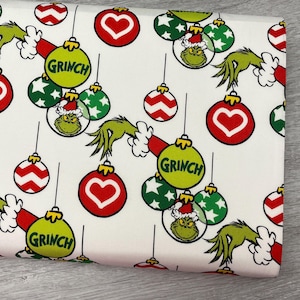 May include: White fabric featuring a repeating pattern of Christmas ornaments. The ornaments include red hearts, green stars, and Grinch faces. The Grinch's hand and the word "Grinch" are also featured.