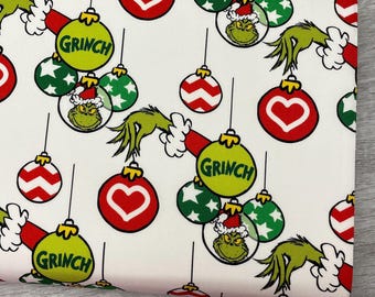 May include: White fabric with a repeating pattern of Christmas ornaments. The ornaments include red hearts, green stars, and Grinch faces. The Grinch's hand and the word "Grinch" are also featured.