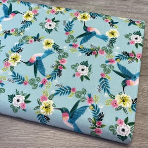 May include: Light blue fabric with a repeating pattern of hummingbirds, flowers, and leaves. The hummingbirds are blue, pink, and yellow, and the flowers are white, pink, and yellow. The leaves are green.