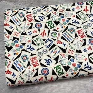 May include: A fabric featuring a repeating pattern of off-white Mahjong tiles against a black backdrop. The tiles display diverse symbols, numbers, and Chinese characters in red, green, blue, and black. The fabric is folded, revealing the pattern's repeating design.
