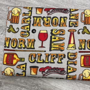 May include: Gray fabric with a repeating pattern of beer mugs, wine glasses, and whiskey bottles. The design includes the words "Cheers", "Norm", "Sam", "Cliff", and "Dia" in a bold, yellow font with red and black accents. An 8-ball is also visible.