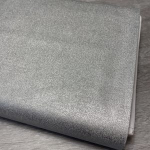 May include: A sheet of silver glitter fabric with a textured surface. The fabric is folded over, revealing a white edge. The material has a sparkly, metallic appearance.