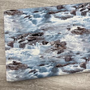 May include: A folded fabric piece displaying a detailed print of a waterfall scene. The design features rushing water, rocky formations, and shades of blue and white, creating a realistic landscape pattern. The edge of the fabric is visible, showing its texture.